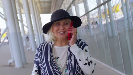 Senior-tourist-grandmother-woman-walking,-international-airport-hall-using-mobile-phone-conversation