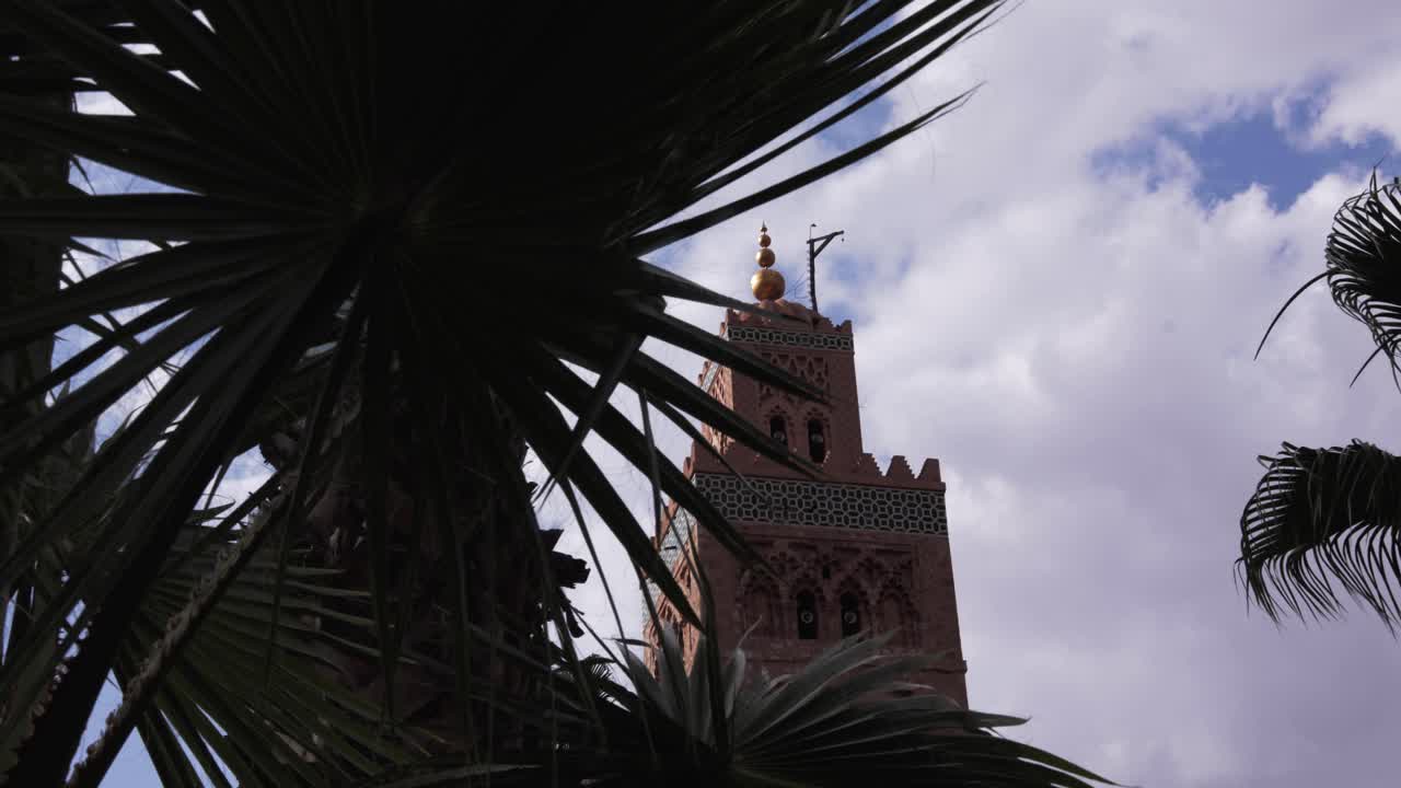 A steady shot of an ancient minaret framed by lush palm leaves &ndash; an iconic view of Marrakesh's historic beauty of a famous mosque in the medina