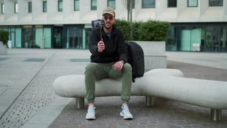 Hombre-Con-Gorra-De-Béisbol-Con-Chaqueta-Negra-Sentado-Fuera-De-La-Abrazadera-Plegable-Conectada-Al-Teléfono-Móvil-Y-Extendiendo-El-Palo-De-Selfie