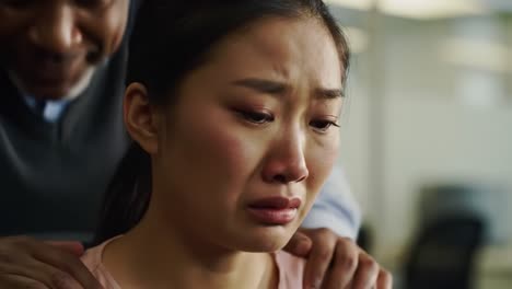 a woman sits at her desk visibly upset, tears streaming down her face. colleagues surround her, unaware of her emotional turmoil. the setting highlights a busy office atmosphere.