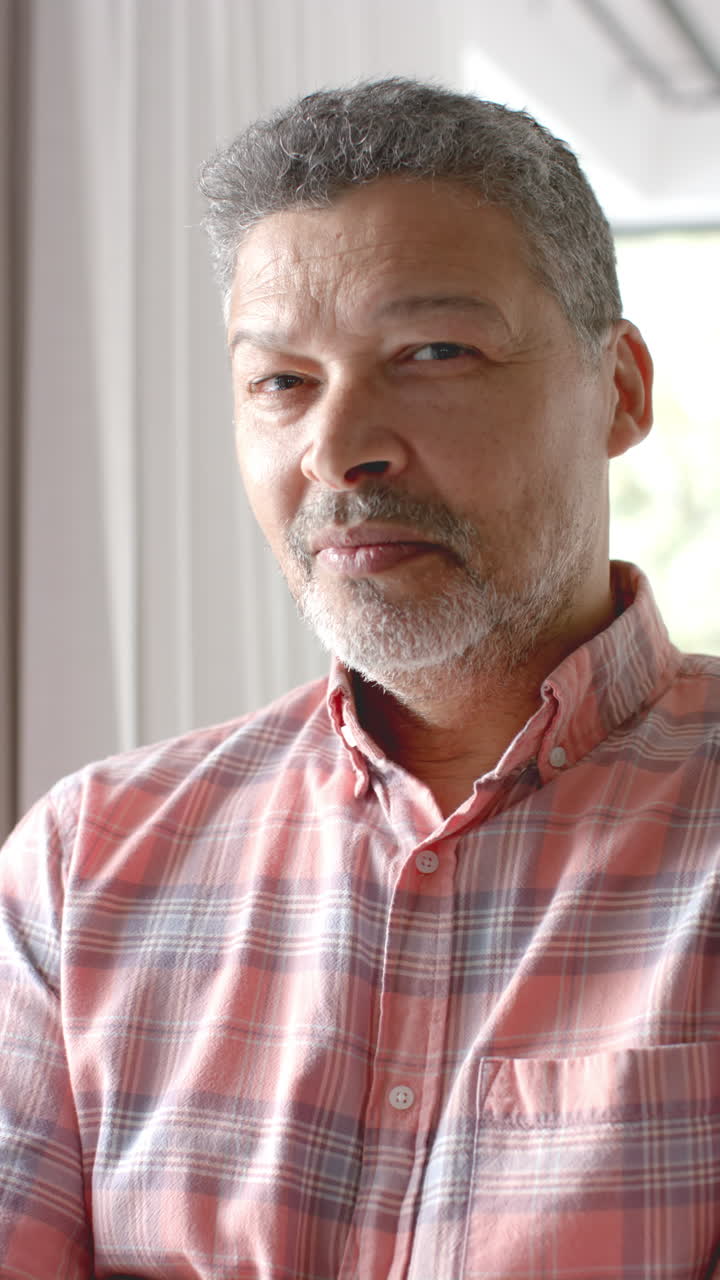 Vertical video portrait of happy senior biracial man smiling in sunny room at home, slow motion