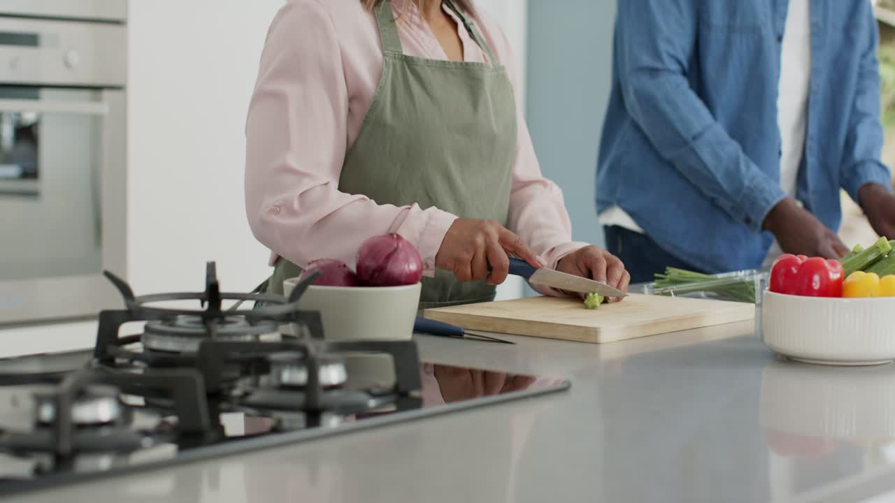 Coppia multietnica che si allunga per tagliare steli su un tagliere con un coltello da chef, cucinando in cucina.