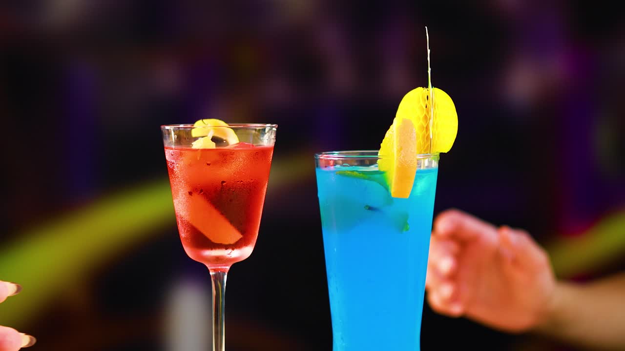 Two hands clink colorful cocktails in a lively bar setting, showcasing vibrant drinks and a celebratory mood