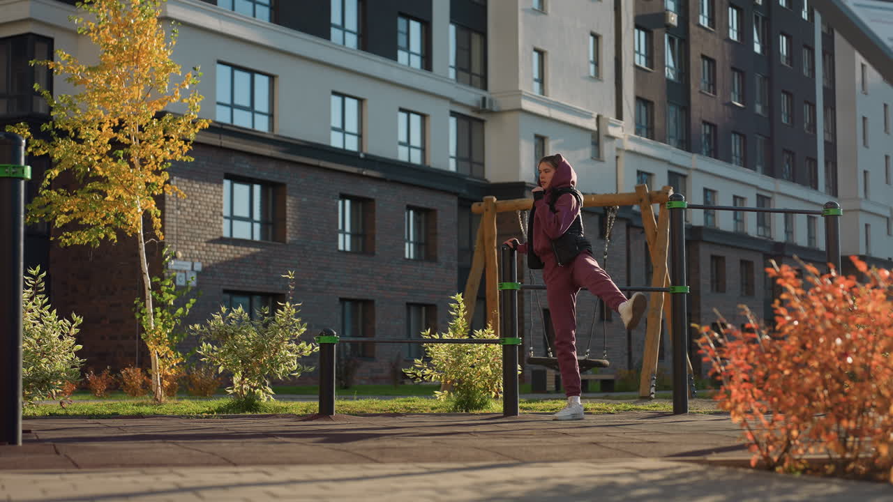 Hoodie wearing athlete gripping metal bar while lifting side leg high during outdoor workout session on fitness park with autumn foliage in background under bright sun and long shadows