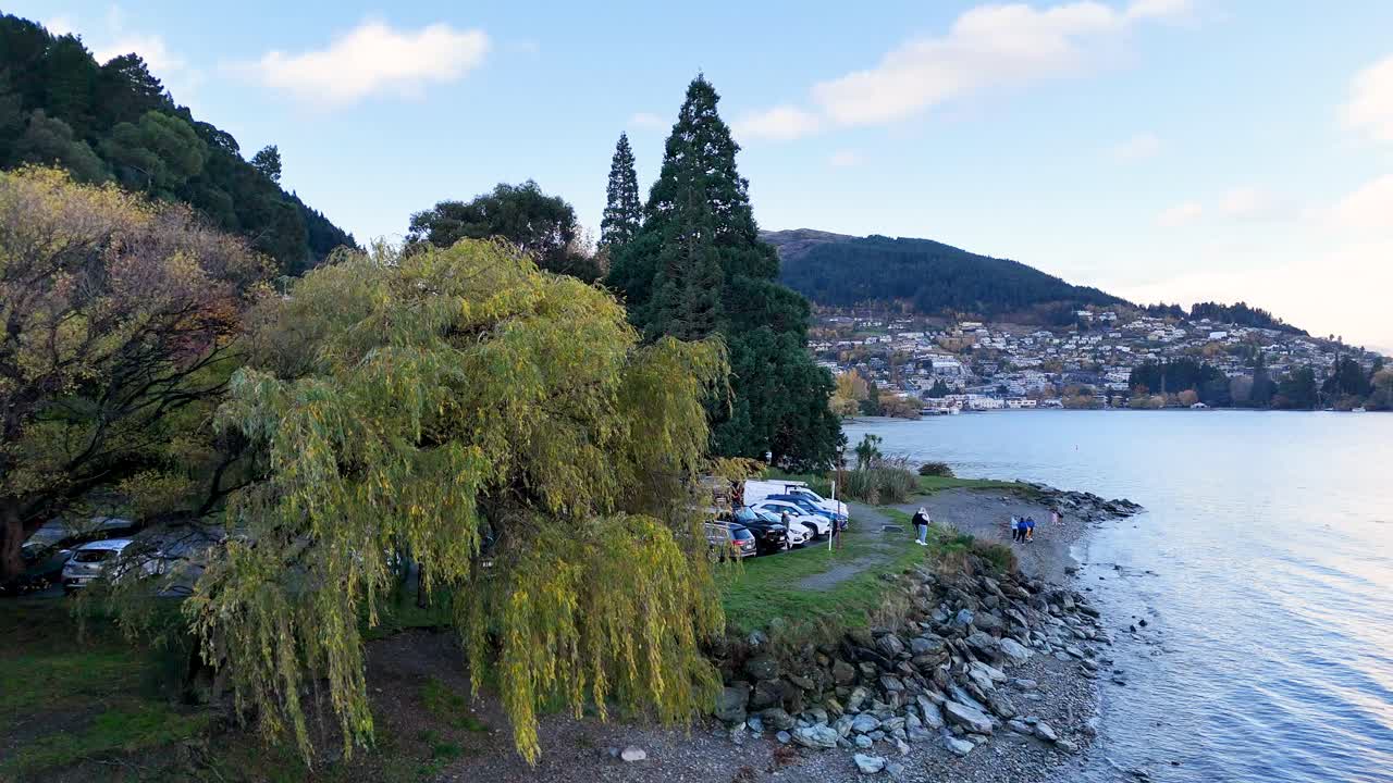 Aerial view of a tranquil riverside park with lush trees, parked cars, and distant mountains under soft daylight