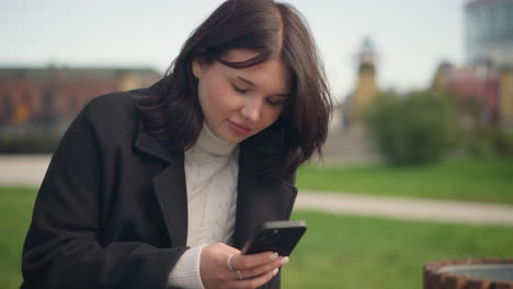 young lady in black coat and white turtleneck focused on her phone with a subtle smile, residential building in the background, sitting outdoors in a peaceful urban park