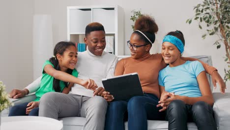 happy family sitting on couch in a newly purchased apartment a married couple and their two daughters browse furniture websites on a tablet, plan a renovation, the girl chooses furnishings