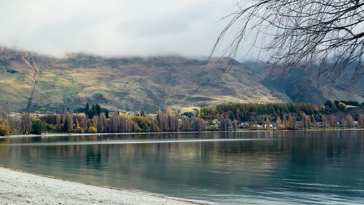 People walk by Lake Wanaka under cloudy skies, surrounded by mountains and trees, creating a peaceful, scenic atmosphere
