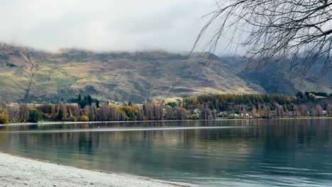 people walk by lake wanaka under cloudy skies, surrounded by mountains and trees, creating a peaceful, scenic atmosphere