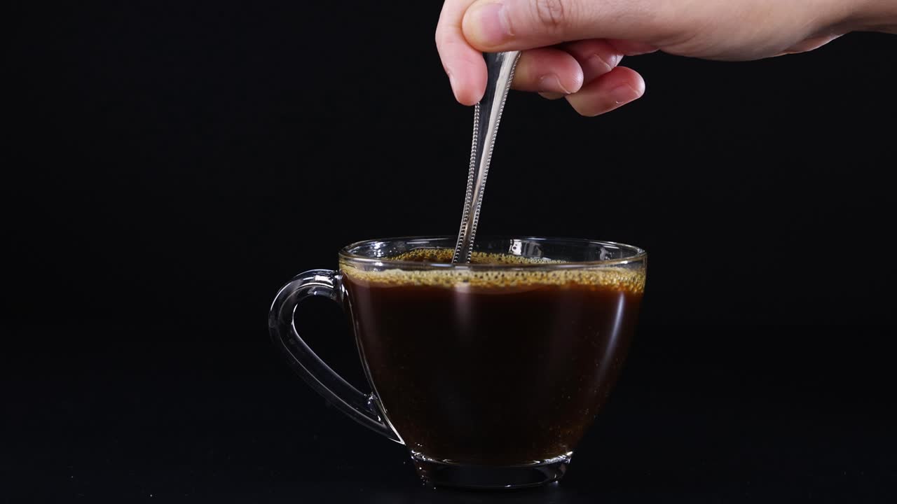A hand stirs coffee in a glass cup against a black background, highlighting motion and simplicity