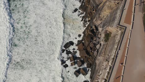 Ocean-waves-hitting-high-cliff-coastline-with-pathway-of-Azenhas-Do-Mar-town,-top-down-view