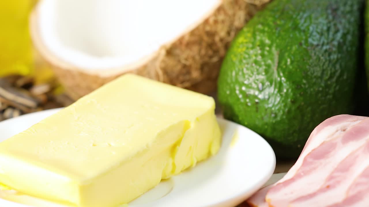 A close-up view of diverse healthy fats including nuts, seeds, butter, avocado, and bacon with bright lighting