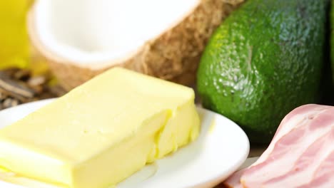 a close-up view of diverse healthy fats including nuts, seeds, butter, avocado, and bacon with bright lighting