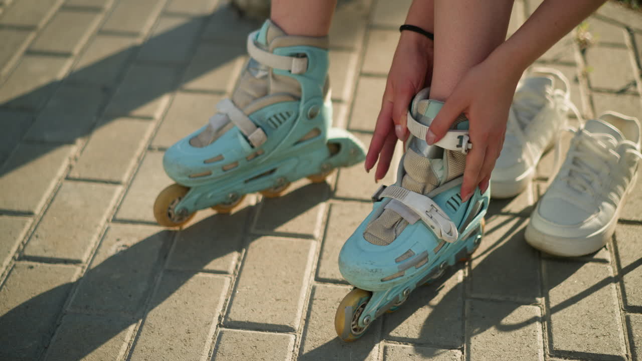 Partial view of individual adjusting roller skate strap on left leg with black wristband under warm sunlight, white sneakers lying nearby on pavement, with greenery softly blurred in background