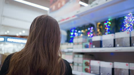 back view of woman browsing through shopping mall, admiring shimmering decorative lights in background, shelves stocked with festive lighting products illuminate well-lit retail space