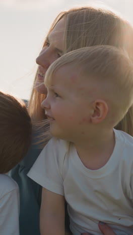a woman in a blue dress sits on the grass, smiling as she takes a picture with a smartphone. her two sons, both wearing white shirts, joyfully join her in capturing this sunny outdoor memory together