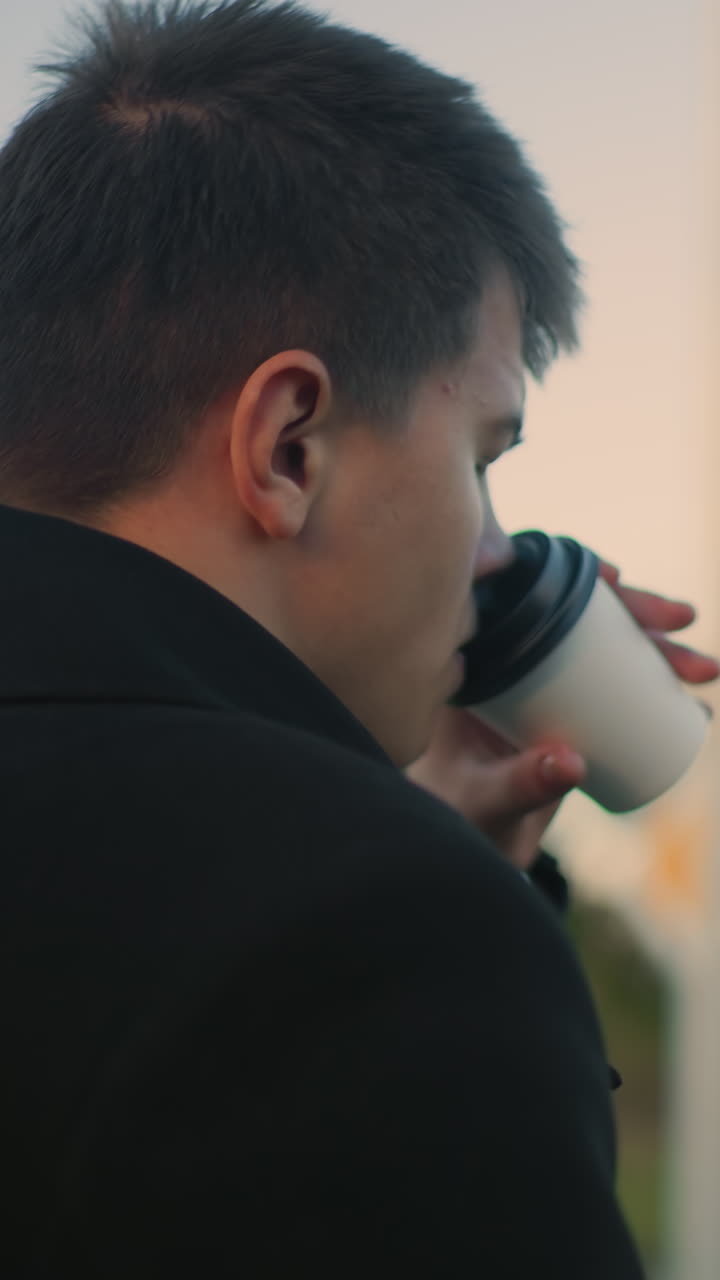 검은 코트를 입은 남자가 차를 마시며 은 조명 아래 녹색과 건물의 흐릿한 배경을 특징으로하는 야외 환경에 깊이 집중합니다.