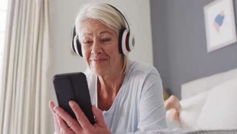 happy senior caucasian woman laying on bed in bedroom, using smartphone and wearing headphones