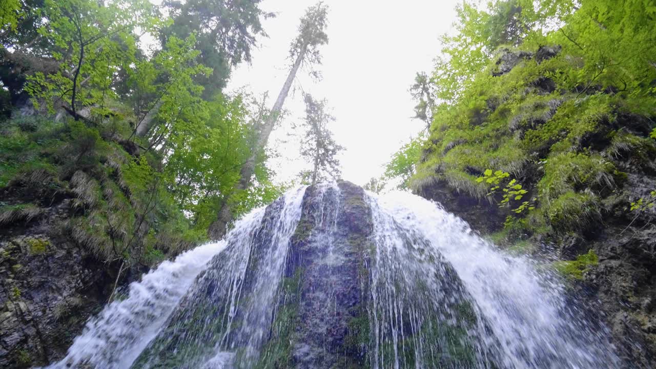 Primo piano di una cascata divisa da una roccia, circondata da rocce nere muschiose, con una panoramica verticale fino al cielo luminoso e soleggiato.