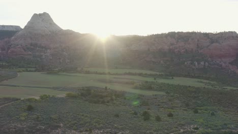 Drone-flies-into-sunset-over-green-grass-valley-with-lens-flare-with-majestic-rocky-cliffs-in-Zion-National-Park