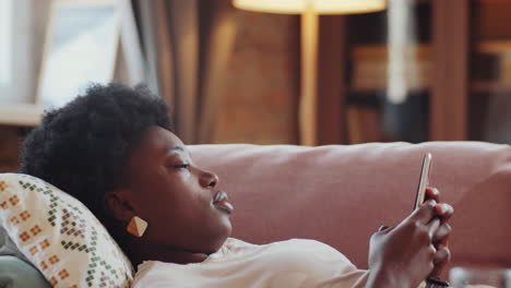 african american woman lying on couch and using smartphone