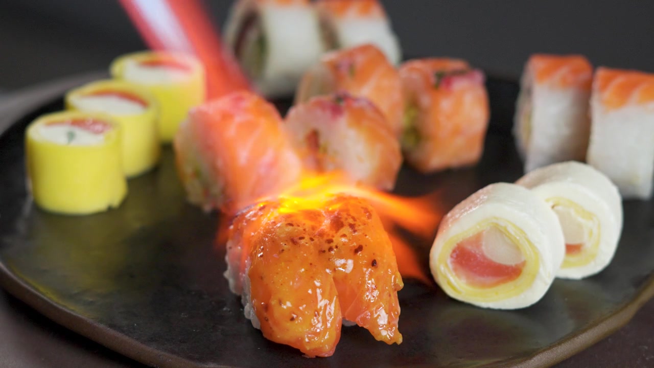 Nahaufnahme eines Kochs, der einen Bunsenbrenner benutzt, um eine Lachs-Sushi-Rolle anzubraten