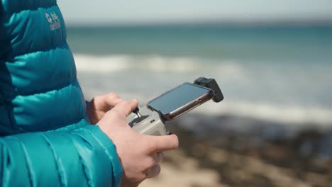 drone pilot remote controlling flying drone while wearing a blue jacket at the beach on a sunny day