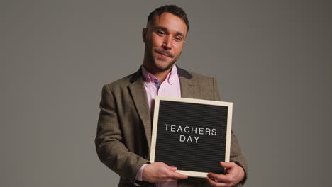 studio portrait of mature male teacher standing against grey background holding notice board reading teachers day