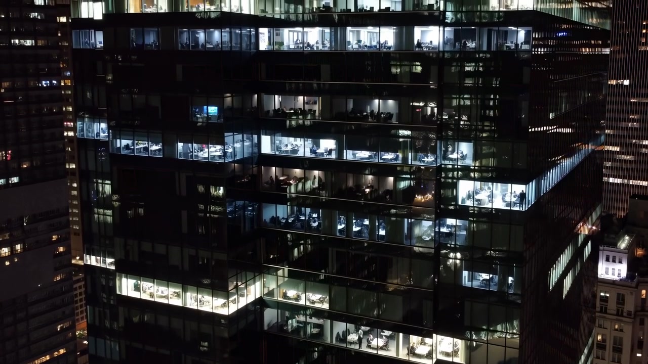 Un tranquillo paesaggio urbano notturno, ripreso da una prospettiva aerea, che mostra grattacieli illuminati da strutture in vetro e metallo. La scena presenta un'atmosfera serena