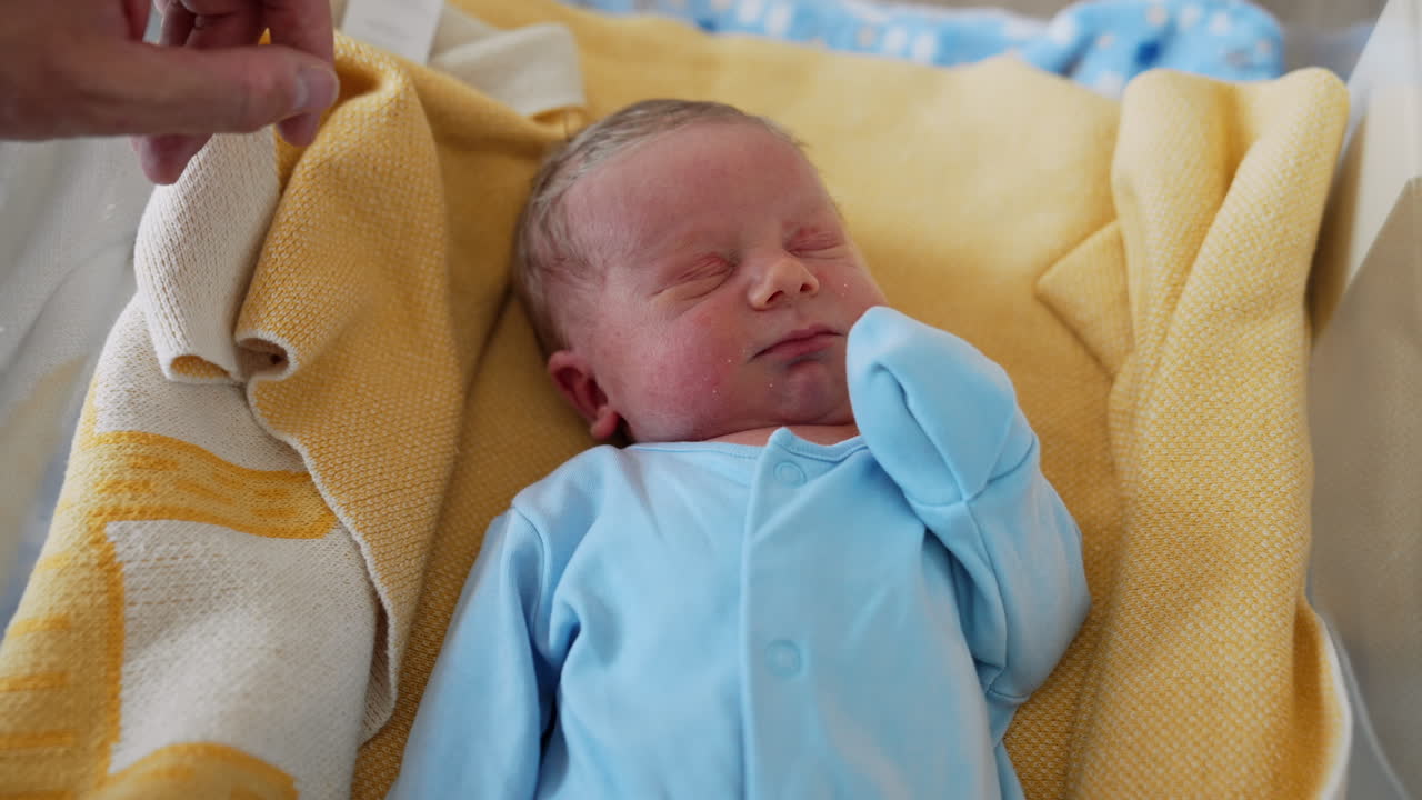 Adorable newborn boy with fair hair. Little baby in a blue bodysuit sleeping after birth in hospital. Close up.