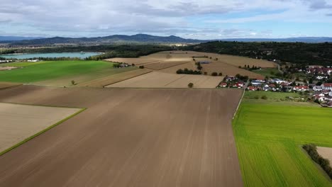 Vuelo-De-Drones-Sobre-Campos-Cosechados-En-Kaiserstuhl-En-Un-Día-De-Verano-Nublado,-Montañas-Y-Estanque-De-Cantera-En-El-Fondo