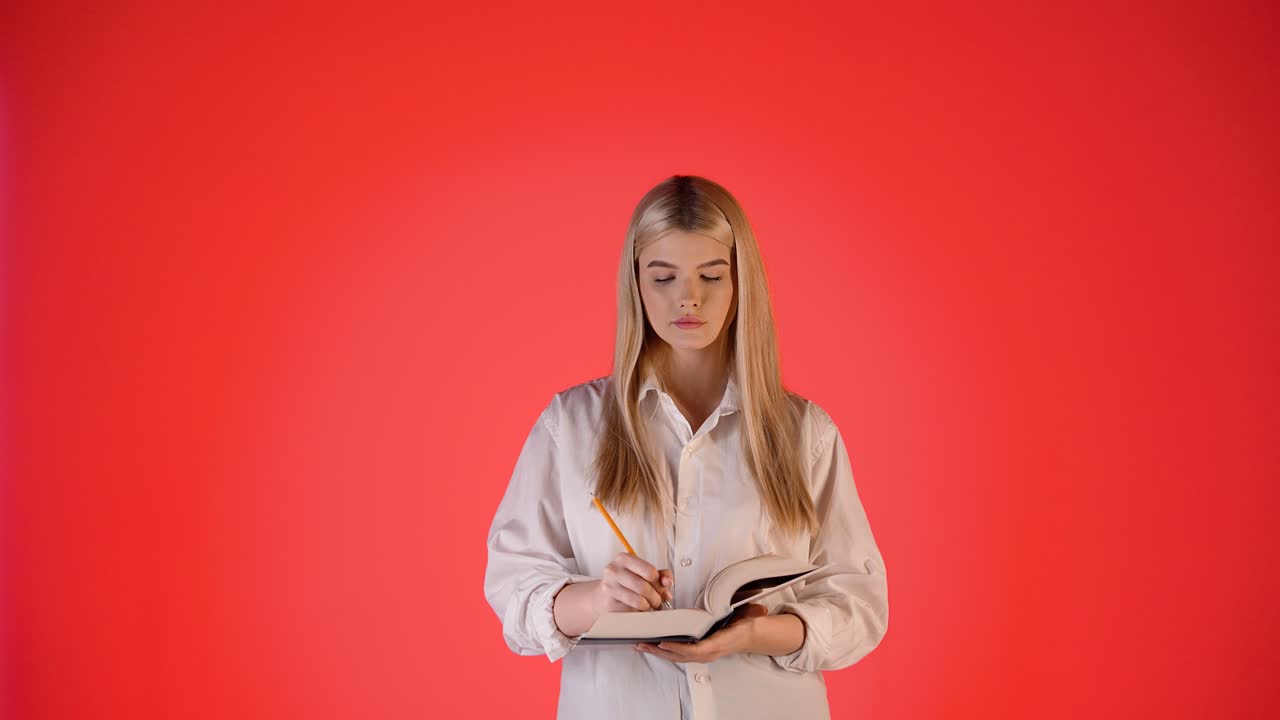 Fokusert ung blond kvinne skriver notater i en papirnotatbok, fargerikt studiobilde med rød bakgrunn.