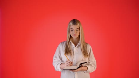 joven rubia enfocada, escribiendo notas en un cuaderno de papel, colorida fotografía de estudio con fondo rojo