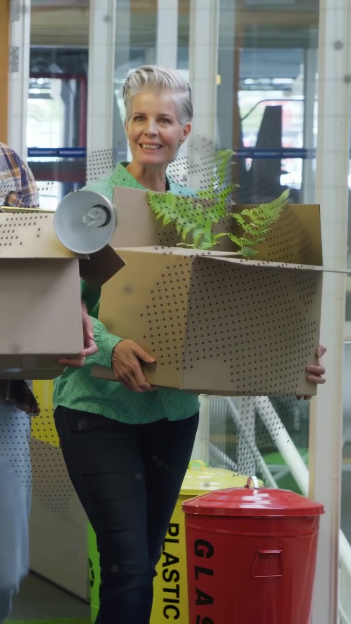 Mature woman entering office with box unpacking fern and lamp organizing workspace with man helping
