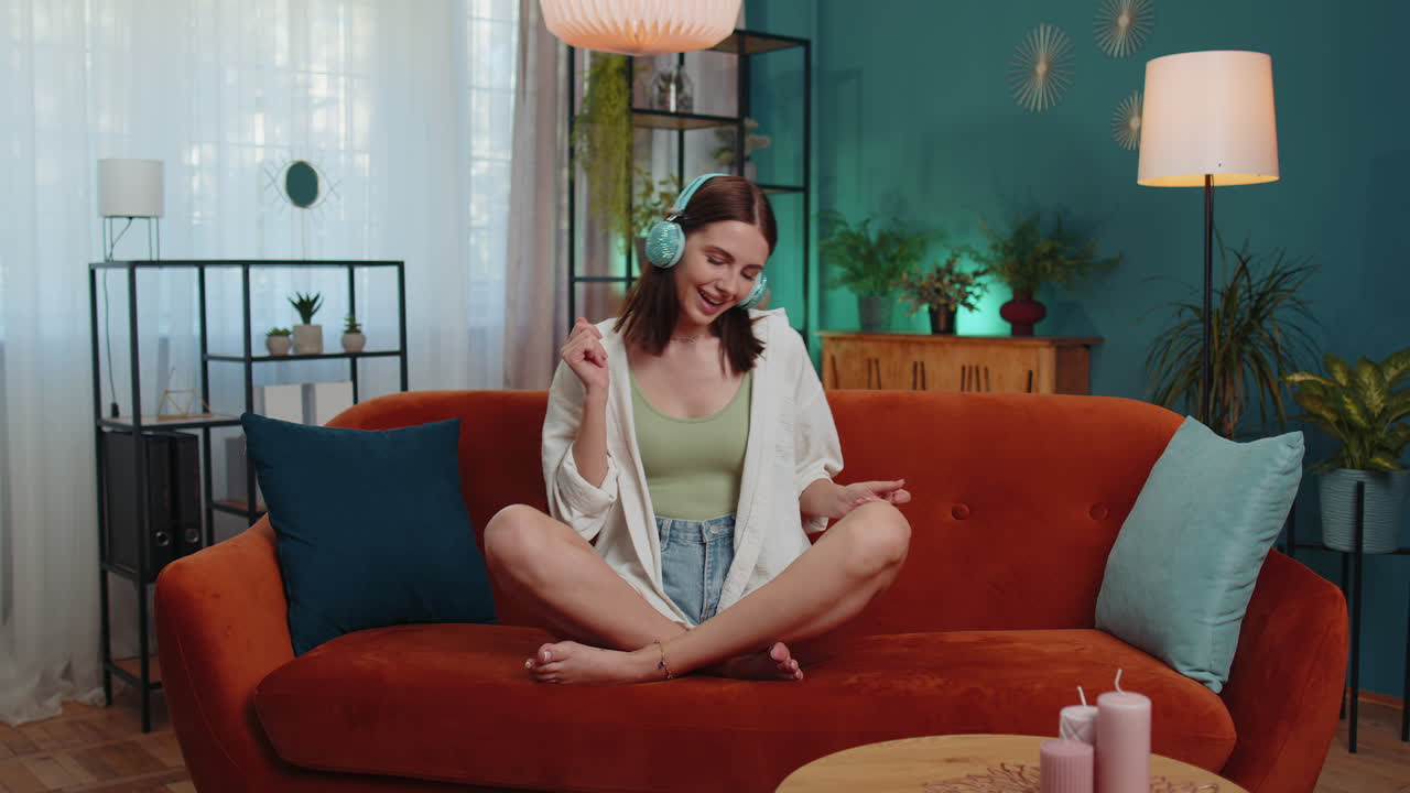 무선 헤드폰을 입은 행복하고 편안한 소녀가 춤을 추고 집 소파에서 좋아하는 록 앤 롤 음악을 듣고 있습니다.