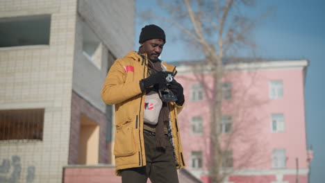 photo maker wearing coat gloves and beanie standing holding camera fixes focus through viewfinder on snowy urban sidewalk under bright winter sun with passersby and colorful buildings in background