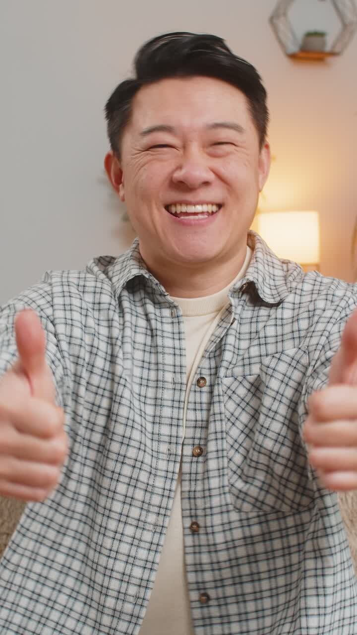 Uomo asiatico seduto sul divano di casa, sorridente e con il pollice in su, felice e soddisfatto.