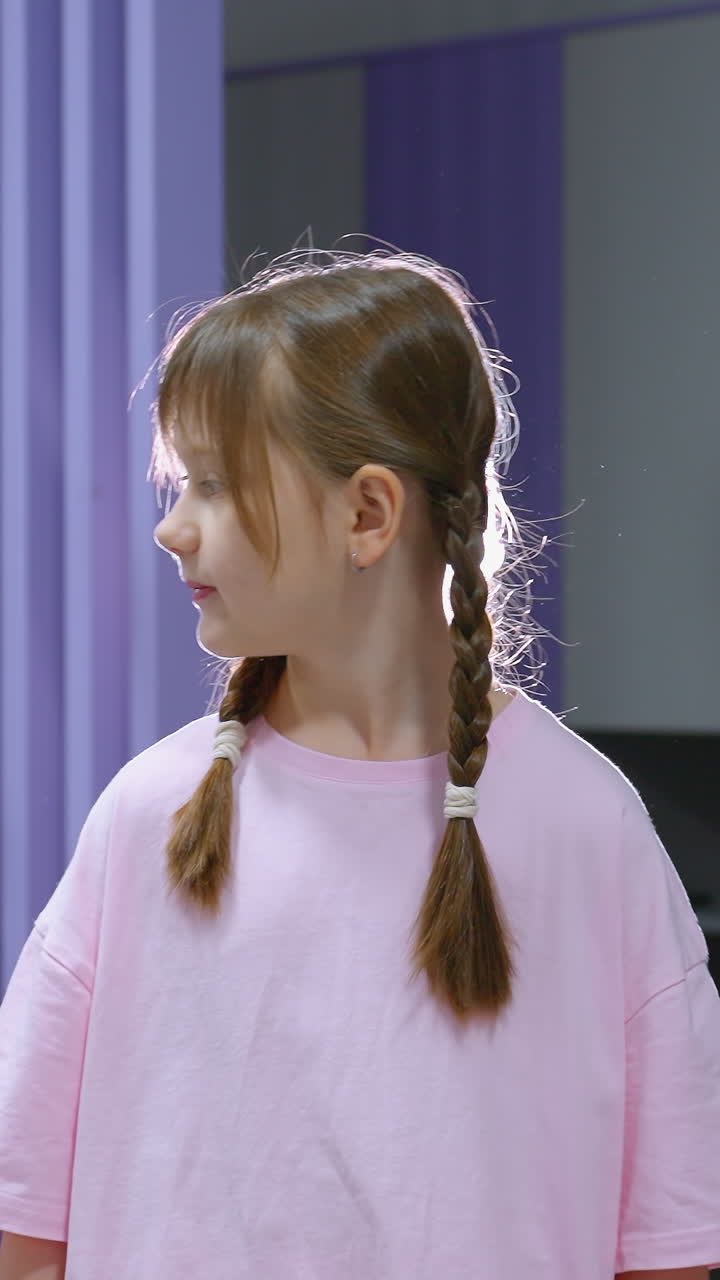 молодая девушка в розовой рубашке с плетеными волосами внимательно смотрит на что-то на заднем плане, корректируя назад, яркий свет освещает позади, оборудование для спортзала видно за ней