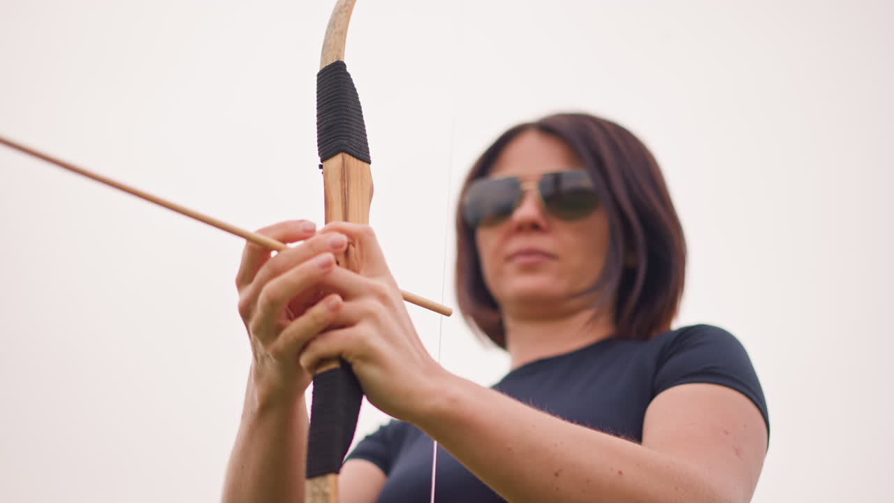 Donna bianca che impugna un arco tradizionale e indossa una gonna con frange, atmosfera da guerriera solitaria in un campo aperto, occhiali da sole, estrazione lenta e ponderata, gonna e cintura in pelle lavorata, mani ferme che preparano la freccia, stile cinematografico attenuato