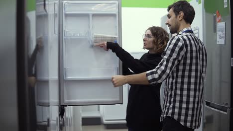 young married couple inspect open door refrigerator, design and quality before buying in a consumer electronics store. discussing characteristics and design