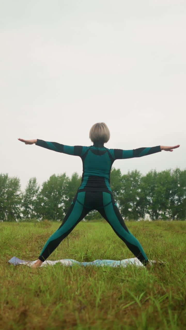 vista trasera de una mujer en traje verde-negro de pie en una alfombra de yoga practicando la postura triangular con los brazos abiertos y las piernas separadas, contra un cielo nublado en un campo tranquilo y cubierto de hierba rodeado de naturaleza
