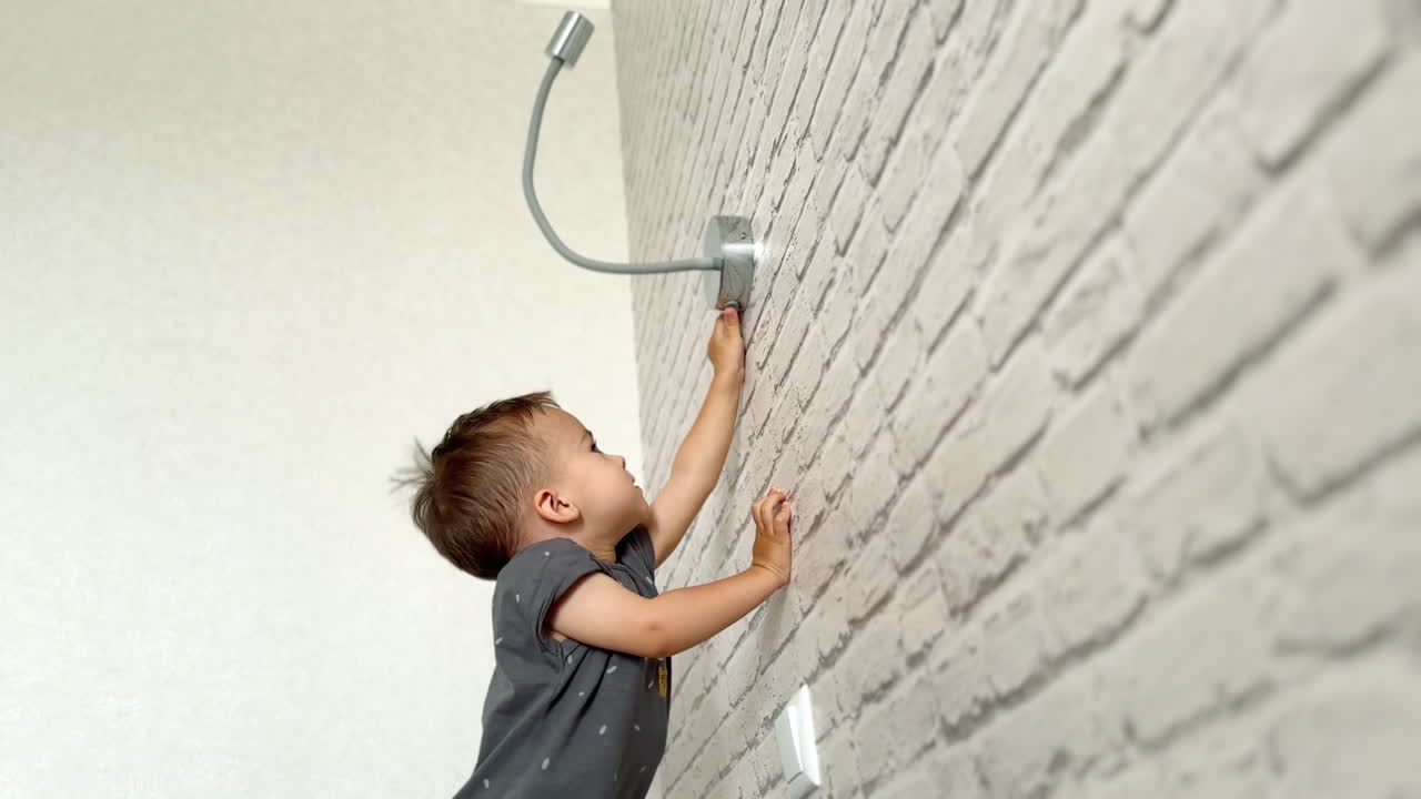 Energetic baby boy wearing grey t-shirt reaching the lamp on the wall. Happy kid pushes the button on the lamp. Low angle view.