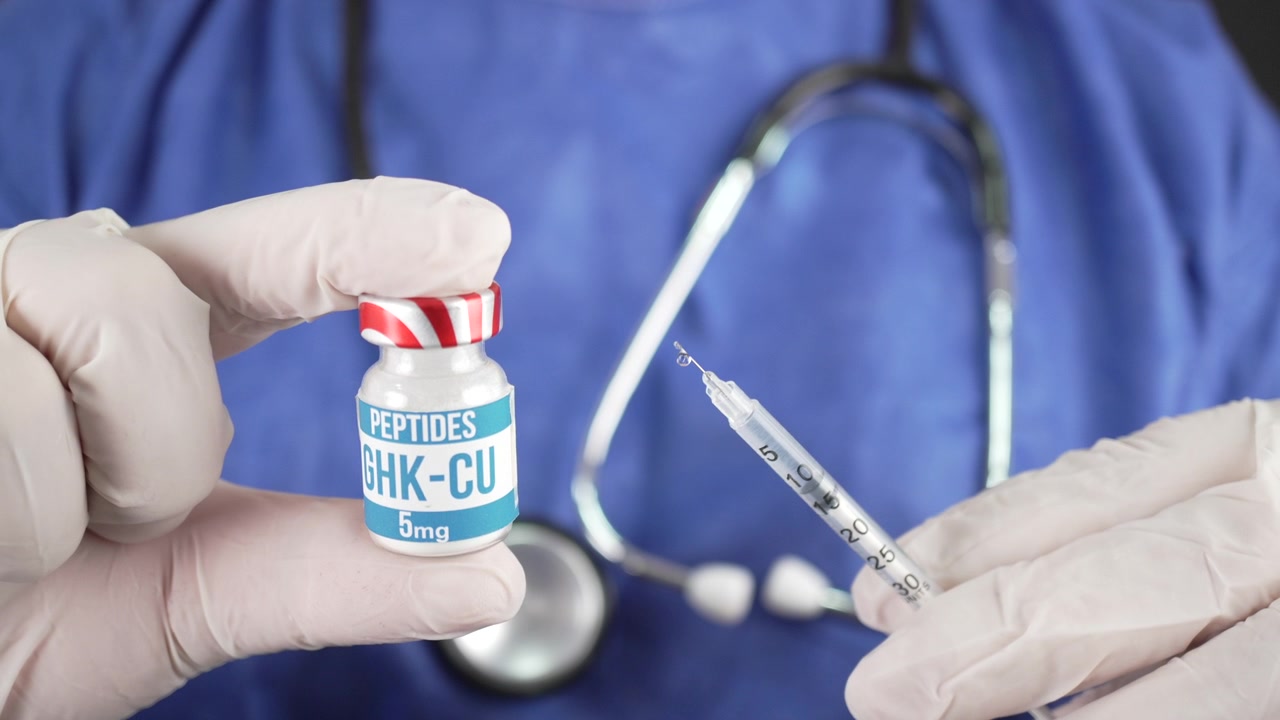 Doctor in latex gloves presents a labeled GHK-CU peptide bottle beside a prepared injection needle against a scrub uniform background.