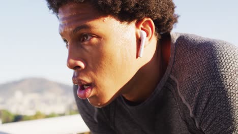 Fit-african-american-man-exercising-outdoors-in-city,-with-wireless-earphones,-resting-on-footbridge