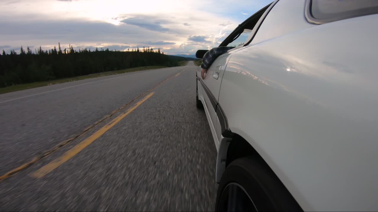 A man taps along to some music while driving his white sports car down the open highway during a colorful sunset.