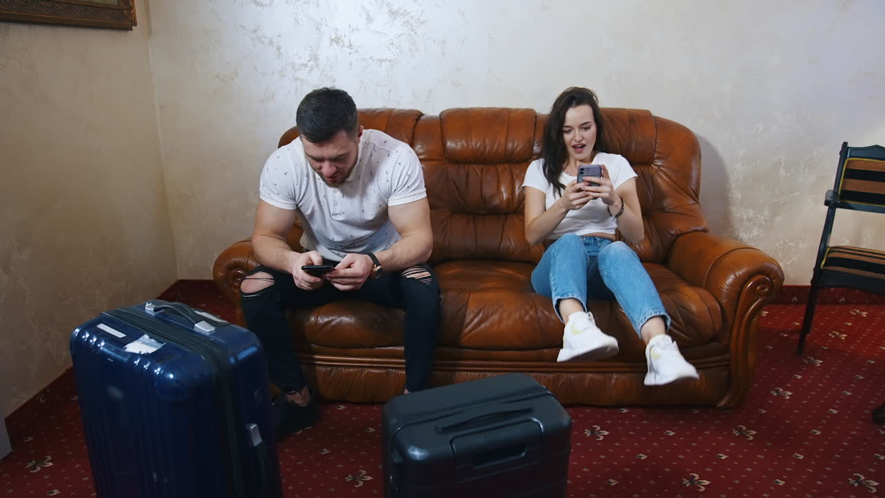 Tourist couple on sofa with travel bags nearby. Young woman and man using phones while waiting for transport at home. Packed suitcases in the hotel.