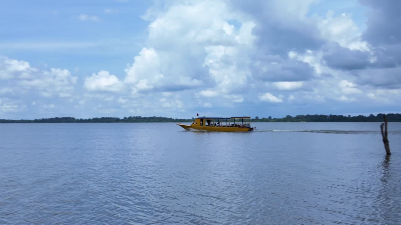 A yellow tourist boat sails on blue water past wooden stakes underneath a blue and cloudy sky on a sunny day