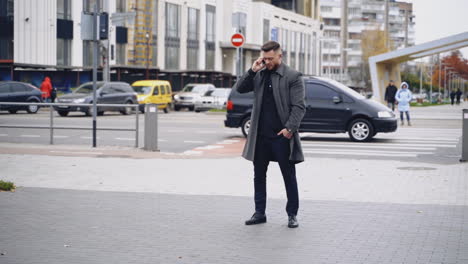 street style. man in an elegant black suit and gray coat. handsome young caucasian man crossing a central road talking on a phone.