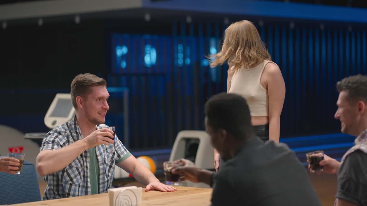 A multi-ethnic group of friends sits at a table together in a bowling alley and talks give five to a joyful man jumping for joy and celebrating victory and a knocked out clash.