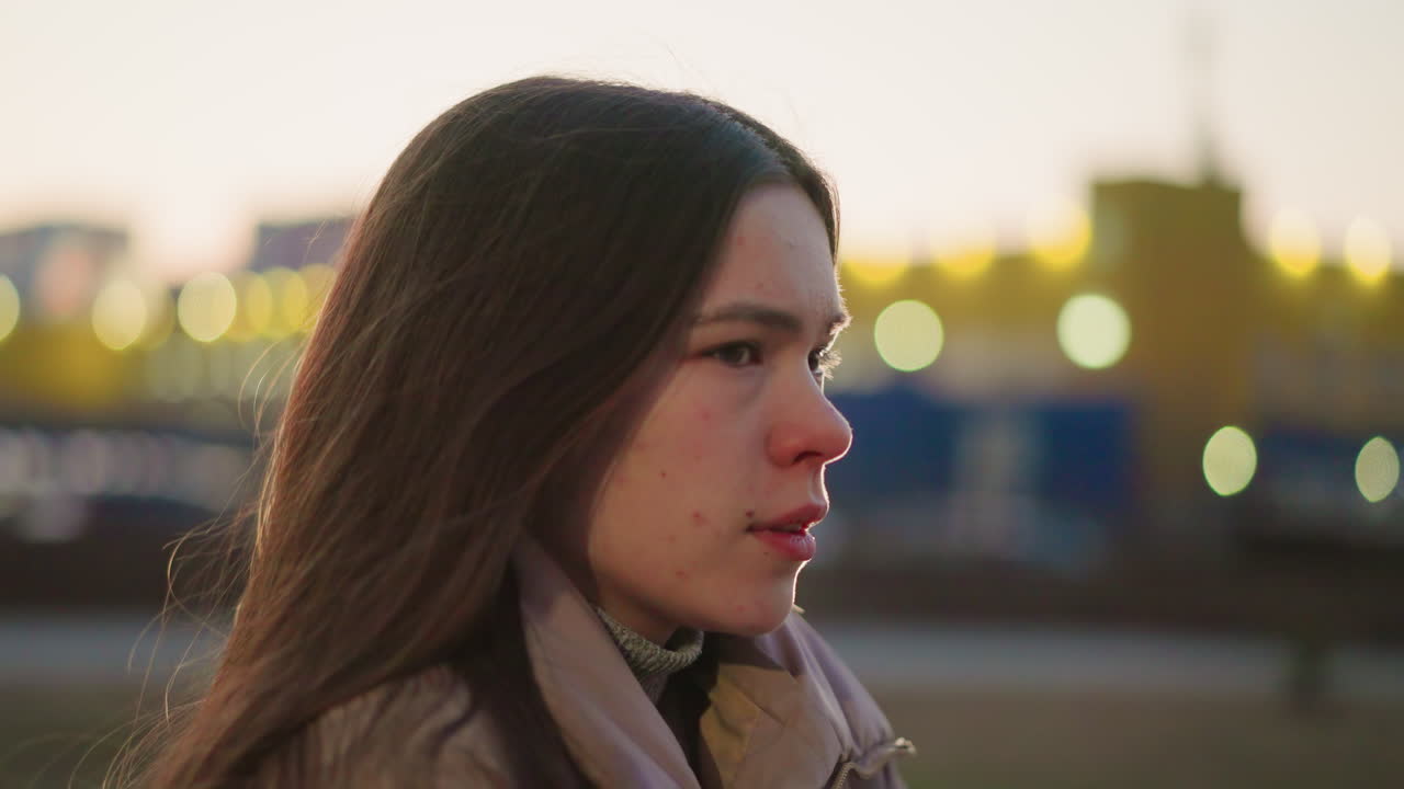 fotografía lateral de una mujer joven con una chaqueta de melocotón, con la boca ligeramente abierta, perdida en sus pensamientos. la suave luz de la tarde y el fondo urbano borroso se suman al estado de ánimo contemplativo